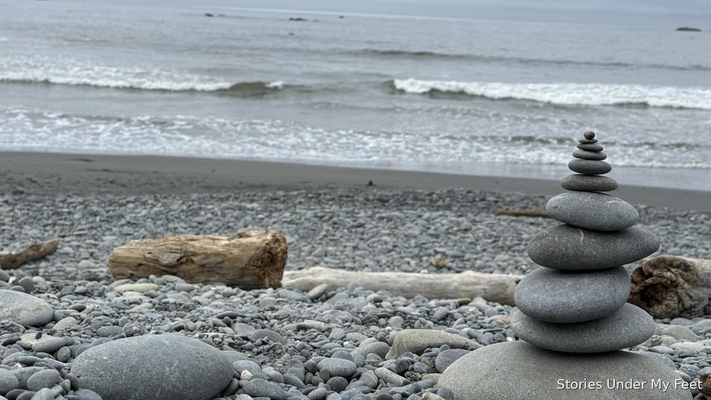 A stone cairn balanced on a driftwood-strewn pebble beach, waves rolling in behind